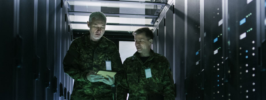 armed service members consulting a handheld computing device while standing in a room of computing equipment and servers